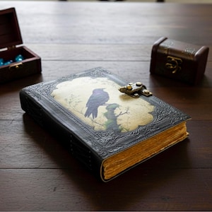 Pu&ograve; includere: Un diario rilegato in pelle nera con un design in rilievo e una chiusura in metallo. La copertina presenta un'illustrazione decorativa di un uccello viola appollaiato su un ramo. Le pagine invecchiate hanno una tonalit&agrave; dorata, suggerendo un'estetica vintage.
