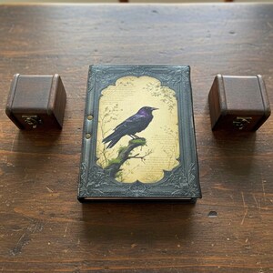 Pu&ograve; includere: Un diario decorativo con un'illustrazione di corvo, affiancato da due piccole scatole di legno. Il diario presenta una copertina nera con una cornice ornata e un'immagine stampata di un corvo dalle sfumature viola appollaiato su un ramo. Le scatole hanno chiusure in metallo.