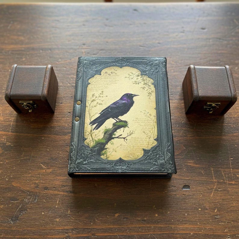 Pu&ograve; includere: Un diario decorativo con un'illustrazione di corvo, affiancato da due piccole scatole di legno. Il diario presenta una copertina nera con una cornice ornata e un'immagine stampata di un corvo dalle sfumature viola appollaiato su un ramo. Le scatole hanno chiusure in metallo.