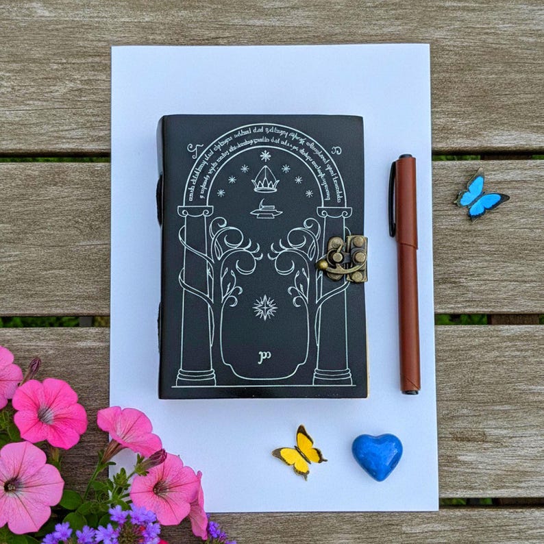 Puede incluir: Un diario negro con un dise&ntilde;o decorativo y cierre, un bol&iacute;grafo marr&oacute;n y mariposas decorativas y un coraz&oacute;n. El diario presenta un dise&ntilde;o blanco de un arco, &aacute;rboles y texto. El bol&iacute;grafo est&aacute; al lado del diario.