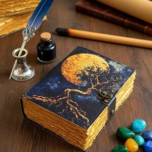 May include: A leather-bound journal with a celestial design featuring a tree and moon. The journal has aged-looking pages and a clasp. A blue feather quill, ink bottle, and gemstones are nearby.