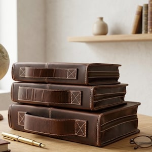 May include: A stack of three rectangular brown leather cases with handles, each featuring a zippered closure and visible white stitching. The cases are arranged in descending order of size, with a gold-toned pen and glasses resting nearby.