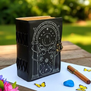 Puede incluir: Diario de cuero negro con dise&ntilde;o celestial blanco y cierre de metal. El diario tiene una columna vertebral cosida y un dise&ntilde;o decorativo en la cubierta frontal. Un bol&iacute;grafo de madera, una piedra azul y decoraciones de mariposas amarillas est&aacute;n cerca.