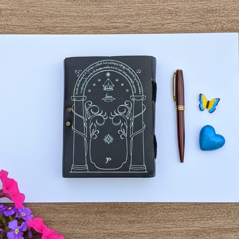Puede incluir: Diario de cuero negro con un dise&ntilde;o plateado en relieve que presenta un arco, estrellas y texto. Un bol&iacute;grafo marr&oacute;n con detalles dorados, un coraz&oacute;n azul y una mariposa est&aacute;n junto al diario. El diario est&aacute; cerrado con una correa.