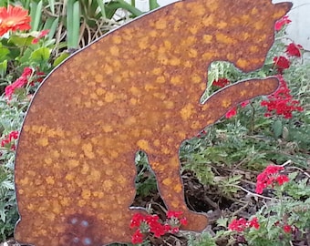 May include: A rusty metal silhouette of a cat sitting in a garden. The cat is brown and orange with a spotted pattern. The cat is surrounded by green plants and red flowers.