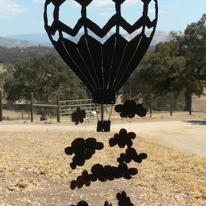 May include: Black metal hot air balloon mobile with cloud accents. The balloon features a geometric design and a basket. The clouds are suspended below the balloon, creating a whimsical outdoor decoration. The background shows a rural landscape.