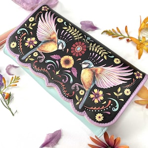 May include: A black wallet with a vibrant bird and floral pattern. The design includes two birds with yellow and blue plumage, set against a backdrop of pink, yellow, and purple flowers. The wallet has a scalloped edge and a pale blue base.