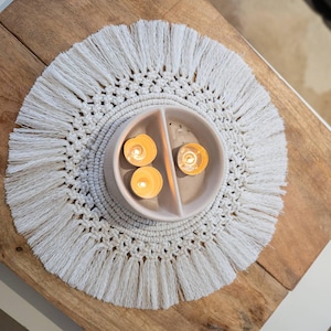 May include: A circular macrame placemat with a fringed edge surrounds a divided ceramic bowl holding three lit candles. The candles emit a warm glow, set on a wooden table.