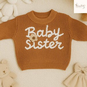 May include: A rust-colored baby sweater with the words "Baby Sister" embroidered in white script. A small flower applique is placed between the words. The sweater has a ribbed knit texture and a round neckline. The sweater is surrounded by baby toys.
