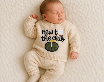 May include: A newborn baby wearing a cream-colored knit sweater and pants set. The sweater features the text "newt the club" with a golf ball and green golf course design. The baby is lying on a cream-colored blanket.