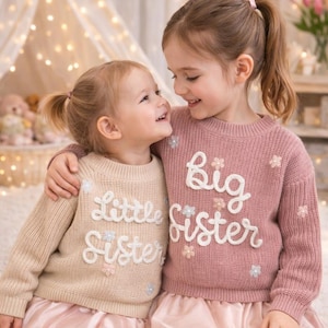 May include: Two long-sleeved knit sweaters, one in beige with the text Little Sister and one in dusty rose with the text Big Sister. Both sweaters feature a ribbed crew neckline, cuffs, and hem, decorated with small, embroidered floral accents.