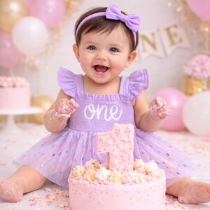 May include: A light purple sleeveless dress with ruffled shoulders and the word one embroidered in white on the chest. The dress features a tulle skirt with colorful sprinkles and is paired with a matching purple headband with a small bow.