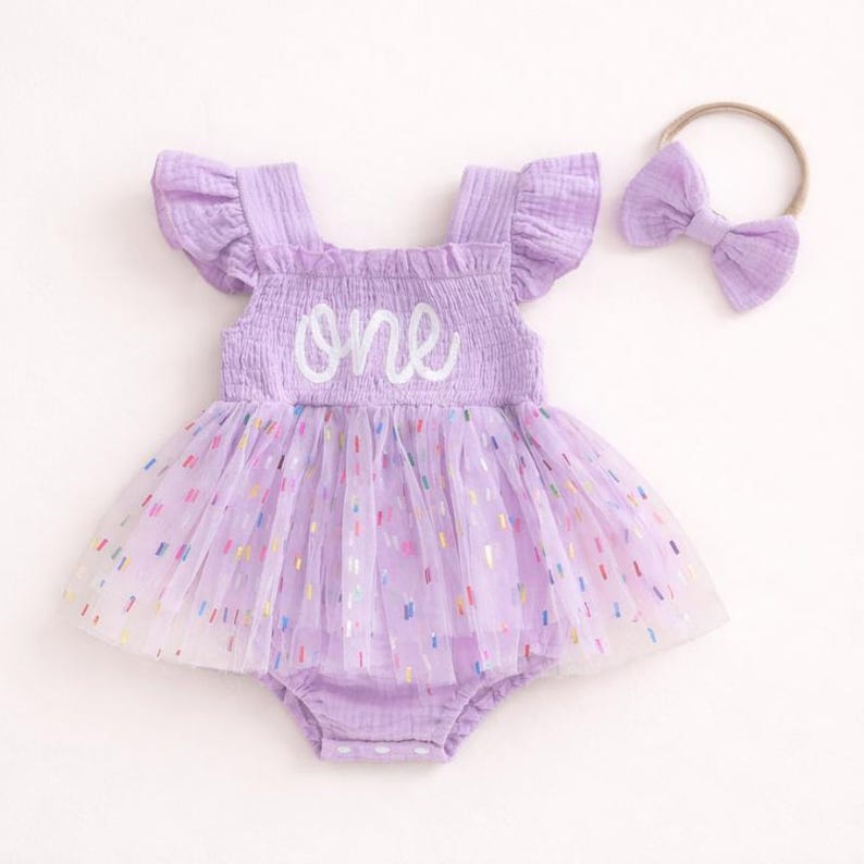 May include: A light purple romper with ruffled sleeves and a smocked bodice featuring the word one in white script. The skirt consists of a layered tulle overlay with multicolored confetti-like accents. A matching purple headband with a bow is included.