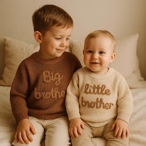 Puede incluir: Dos niños con jerséis de punto a juego. Uno es marrón con "Big brother" cosido en la parte delantera, el otro crema con "Little brother". Ambos jerséis tienen una textura acanalada. Los niños están juntos.