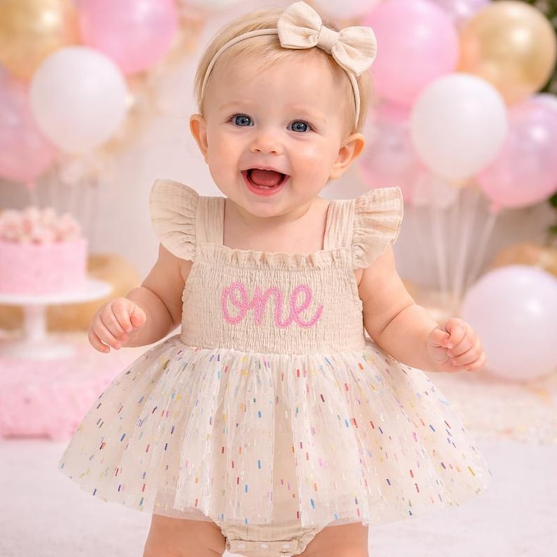 May include: A beige sleeveless romper with ruffled shoulders and a tulle skirt featuring colorful confetti-like flecks. The bodice has a smocked texture with the word one embroidered in pink script. A matching beige headband with a bow is included.