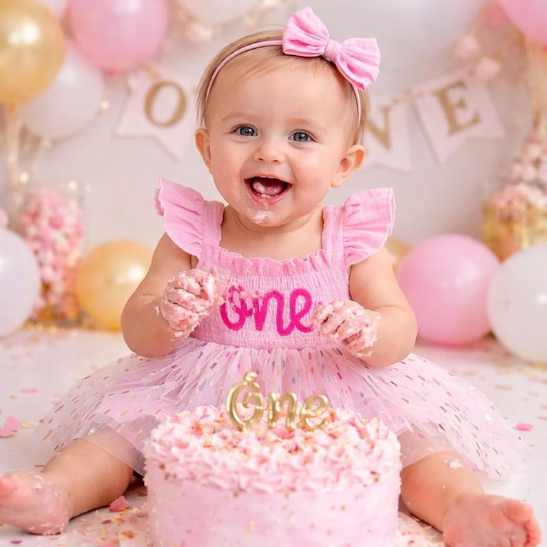 May include: A light pink sleeveless dress with ruffled shoulders and the word one embroidered in dark pink on the bodice. The dress features a tulle skirt with small colorful flecks. A matching pink headband with a bow is worn on the head.