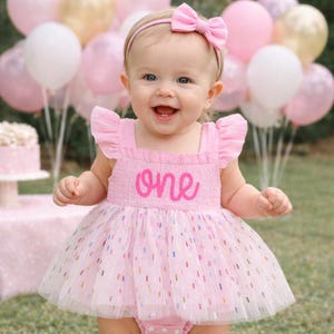 May include: A light pink sleeveless dress with a smocked bodice featuring the word one in cursive script and a tulle skirt with colorful confetti-like accents. A matching pink headband with a bow is worn on the head to complete the outfit.