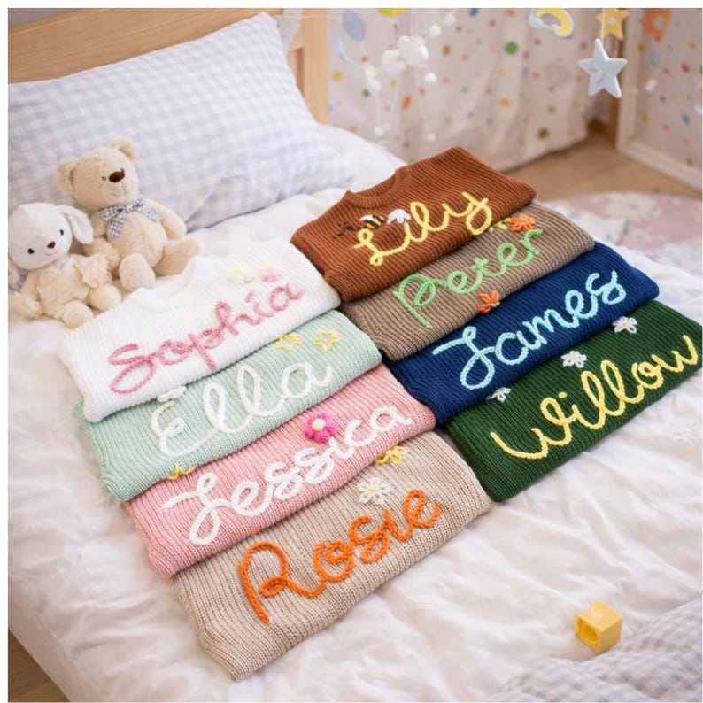 May include: A stack of personalized knit sweaters in various colors, each embroidered with a name in a cursive font. The sweaters are in shades of white, brown, green, pink, and blue. Some sweaters have floral embellishments. A teddy bear and a stuffed bunny are in the background.