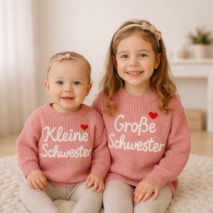 May include: Two young girls wearing matching pink sweaters with white lettering and a red heart. The sweater on the left says "Kleine Schwester" and the sweater on the right says "Große Schwester". Both girls are smiling and wearing headbands.