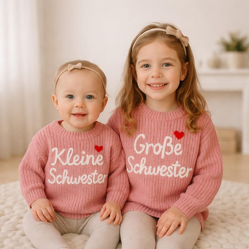 May include: Two young girls wearing matching pink sweaters with white lettering and a red heart. The sweater on the left says "Kleine Schwester" and the sweater on the right says "Gro&szlig;e Schwester". Both girls are smiling and wearing headbands.