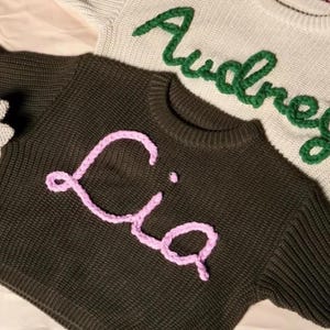 May include: Two knitted sweaters. One is dark green with the name "Lia" in pink yarn. The other is light beige with the name "Audrey" in green yarn. Both sweaters have a crew neck.
