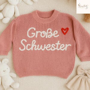 May include: A pink knit sweater with the words "Gro&szlig;e Schwester" embroidered in white, and a red heart. The sweater has a ribbed crew neck and long sleeves. The sweater is likely for a child.