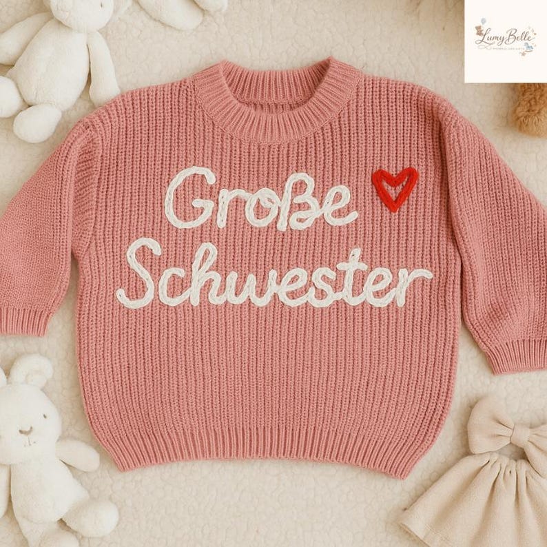 May include: A pink knit sweater with the words "Gro&szlig;e Schwester" embroidered in white, and a red heart. The sweater has a ribbed crew neck and long sleeves. The sweater is likely for a child.