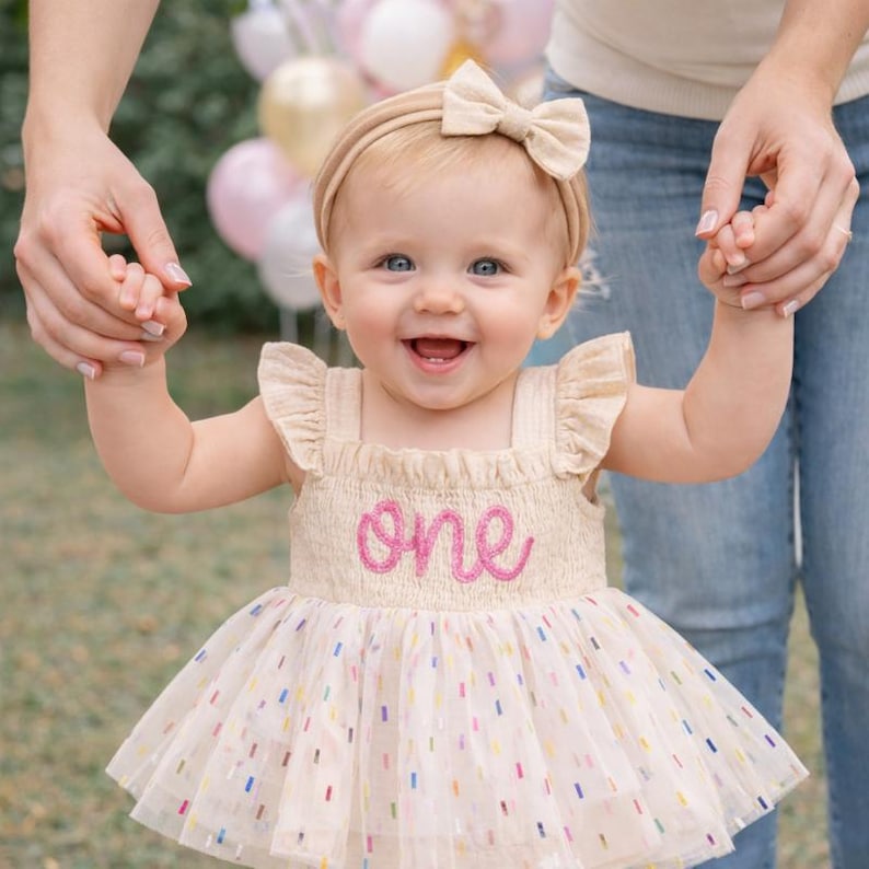 May include: A beige sleeveless dress with ruffled shoulders and a smocked bodice featuring the word one embroidered in pink script. The skirt is made of layered tulle with a colorful confetti pattern. A matching beige headband with a bow is included.