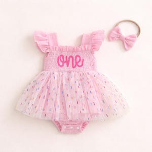 May include: A light pink sleeveless romper with ruffled shoulders and a smocked bodice featuring the word one in dark pink script. The skirt is made of layered tulle with small multicolored confetti-like dashes and includes a matching headband.