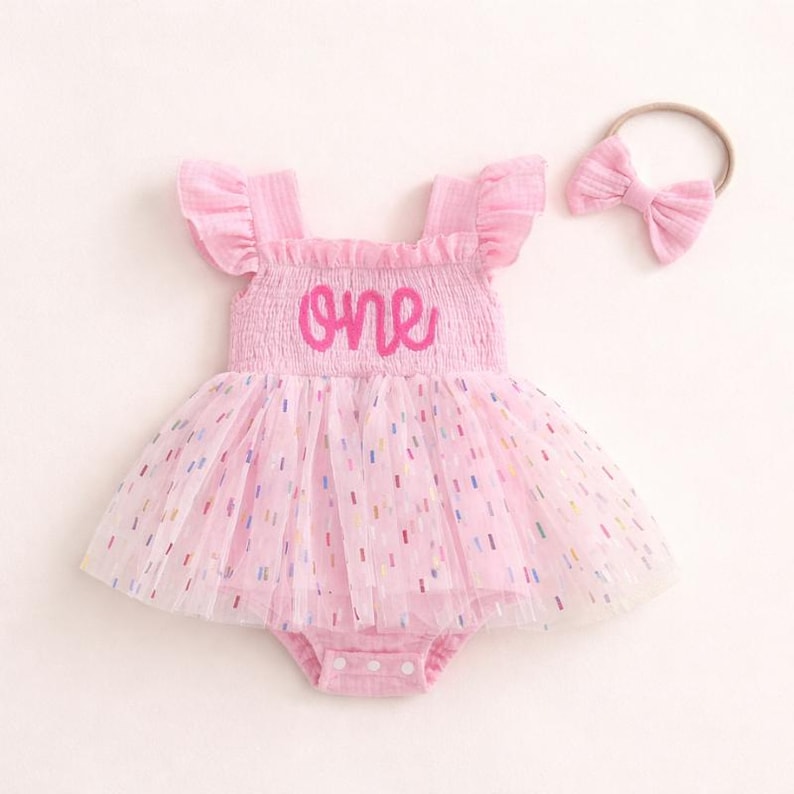 May include: A light pink sleeveless romper with ruffled shoulders and a smocked bodice featuring the word one in dark pink script. The skirt is made of layered tulle with small multicolored confetti-like dashes and includes a matching headband.
