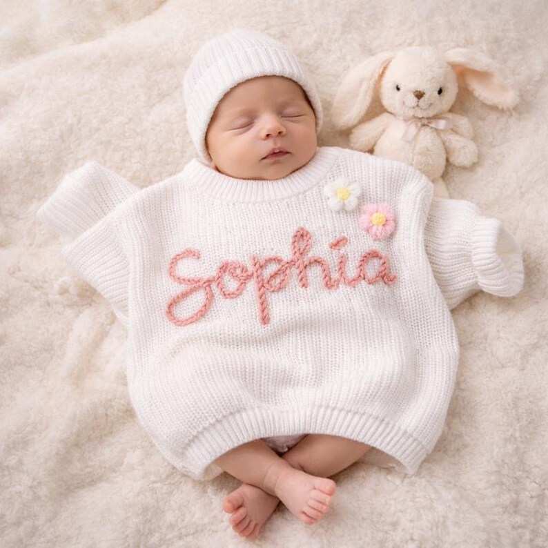 May include: A newborn baby wearing a white knit sweater and matching beanie. The sweater features the name "Sophia" in pink script, along with two small flower appliqu&eacute;s. A plush bunny toy rests beside the baby on a soft, cream-colored blanket.