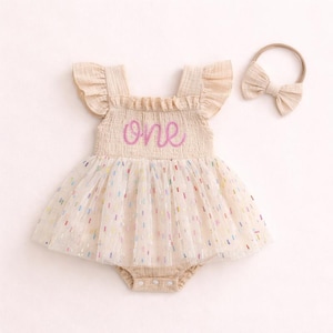 May include: A beige sleeveless romper with ruffled shoulders and a smocked bodice featuring the word one embroidered in pink script. The skirt is made of a sheer tulle overlay with colorful confetti-like flecks and includes a matching bow headband.