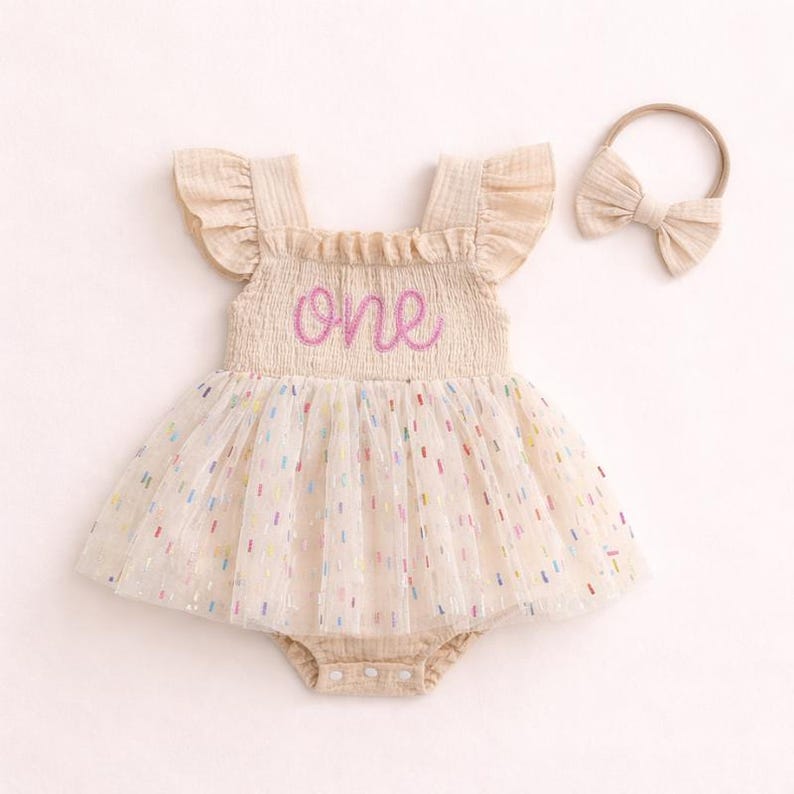 May include: A beige sleeveless romper with ruffled shoulders and a smocked bodice featuring the word one embroidered in pink script. The skirt is made of a sheer tulle overlay with colorful confetti-like flecks and includes a matching bow headband.