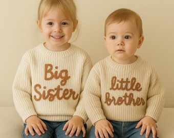May include: Two children wearing cream-colored, long-sleeved, ribbed knit sweaters paired with blue jeans. The left sweater features the brown embroidered text "Big Sister," while the right sweater features the brown embroidered text "little brother."