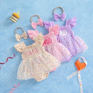 May include: Three infant rompers with ruffled sleeves and tulle skirts featuring colorful confetti dots. Each romper has the word one embroidered on the bodice. Three matching headbands with bows are included, along with a small party hat.