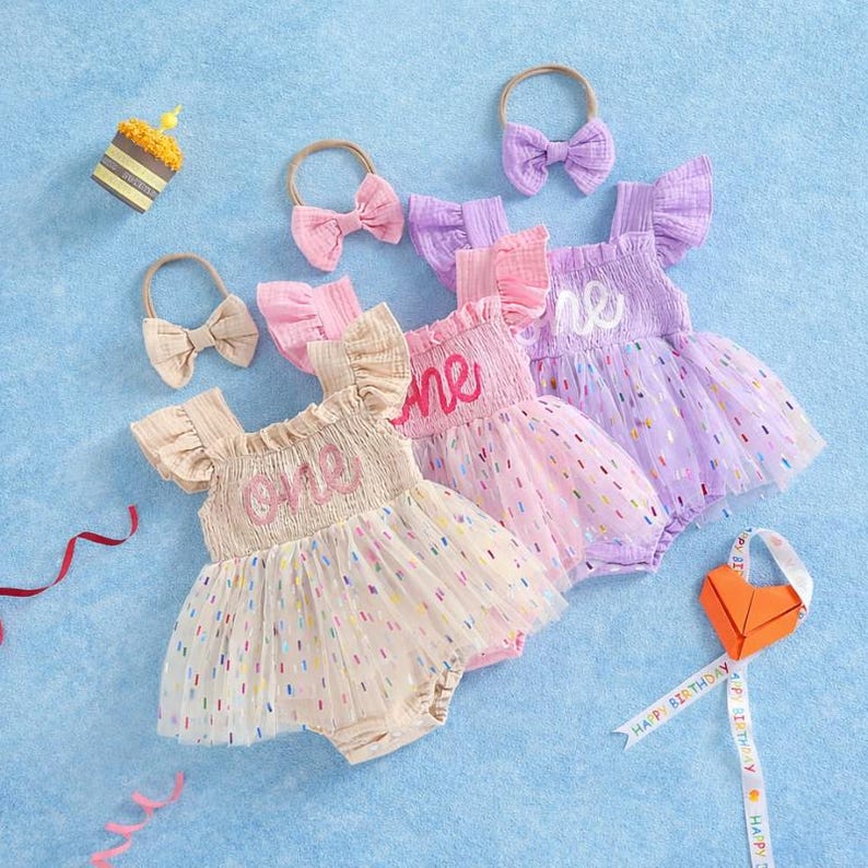 May include: Three infant rompers with ruffled sleeves and tulle skirts featuring colorful confetti dots. Each romper has the word one embroidered on the bodice. Three matching headbands with bows are included, along with a small party hat.