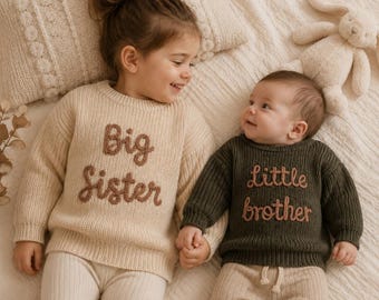 May include: A beige knit sweater with the text Big Sister in brown embroidery and a dark olive green knit sweater with the text little brother in brown embroidery, both featuring long sleeves and a crew neckline.
