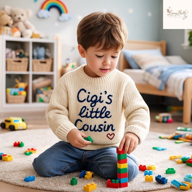 May include: A young child wearing a cream-colored sweater with the embroidered text "Cigi's little Cousin" and a small red heart. The child is playing with colorful building blocks on a neutral-toned rug. The room has a toy car and a rainbow decoration.