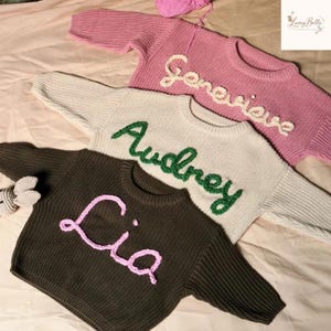 May include: Three personalized knit sweaters in pink, cream, and dark green. Each sweater features a name embroidered in a contrasting color: "Genevieve" in white, "Audrey" in green, and "Lia" in pink. A ball of pink yarn is visible.