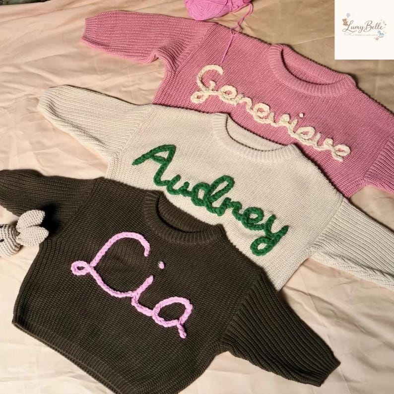 May include: Three personalized knit sweaters in pink, cream, and dark green. Each sweater features a name embroidered in a contrasting color: "Genevieve" in white, "Audrey" in green, and "Lia" in pink. A ball of pink yarn is visible.