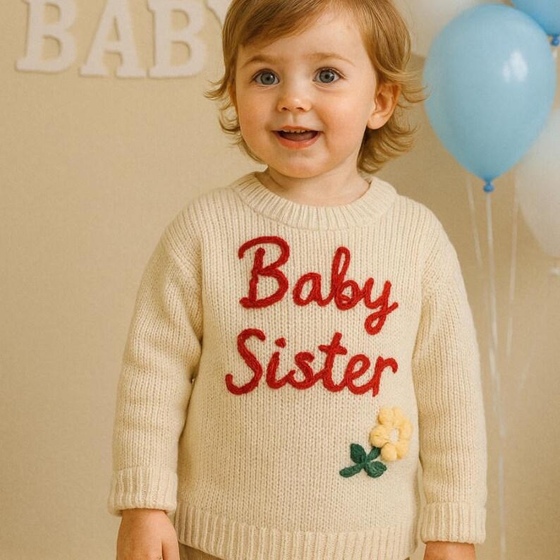 May include: A cream-colored sweater with the words "Baby Sister" embroidered in red. The sweater features a yellow flower and green leaves applique. The background includes a child, balloons, and the word "BABY" in white letters.