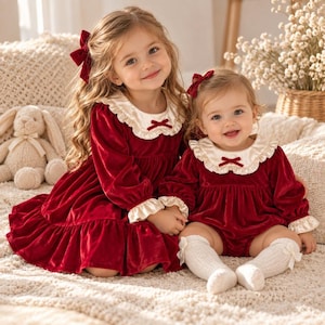 Peut inclure: Deux tenues assorties en velours rouge profond avec des cols à volants couleur crème ornés de petits nœuds rouges. L'une est une robe à manches longues avec un ourlet volanté, l'autre une barboteuse à manches longues avec des chaussettes en tricot.