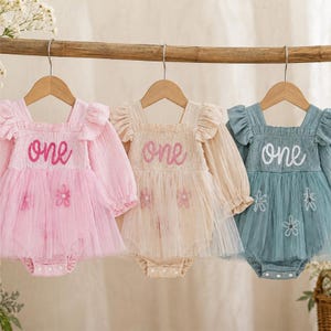 May include: Three long-sleeve rompers hanging on wooden hangers. The rompers are pink, beige, and blue, each featuring a smocked bodice with the word one embroidered on the front, ruffled shoulders, and a tulle skirt overlay with flower appliques.