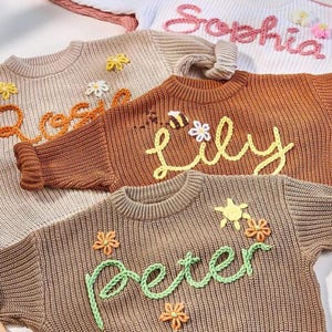 May include: Close-up of several children's sweaters in various colors, each with a name embroidered on the front. The sweaters are adorned with floral and bee embellishments. The names "Sophia", "Lily", "Peter", and "Rose" are visible.