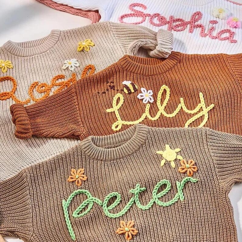 May include: Close-up of several children's sweaters in various colors, each with a name embroidered on the front. The sweaters are adorned with floral and bee embellishments. The names "Sophia", "Lily", "Peter", and "Rose" are visible.