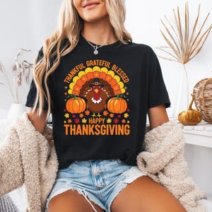 Könnte beinhalten: Schwarzes T-Shirt mit einem Thanksgiving-Motiv. Das Design zeigt einen Truthahn mit einem Pilgerhut, zwei orangefarbene Kürbisse und die Worte "Thankful, Grateful, Blessed" und "Happy Thanksgiving" in Orange und Gelb.