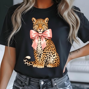 May include: Black t-shirt with a leopard design wearing a pink bow. The leopard has a brown and tan spotted pattern. The bow is a soft pink colour. The t-shirt is a crew neck style.