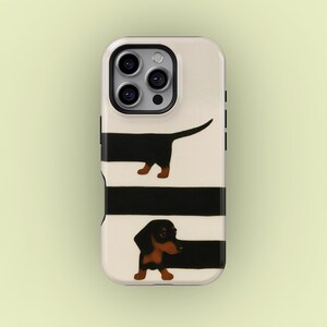 May include: A phone case featuring a cream background with black horizontal stripes and two dachshund illustrations. The dachshunds are black with brown paws and ears. The phone case has a camera cutout and black side buttons.