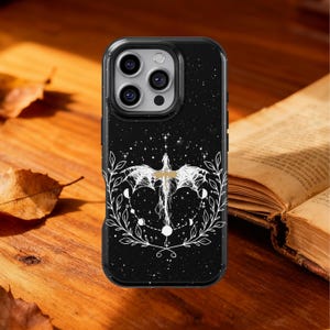 Puede incluir: Funda de teléfono negra con un diseño de dragón blanco, rodeada por una corona floral y elementos celestiales. La funda tiene un fondo estrellado y una cruz dorada. La funda está sobre una superficie de madera.