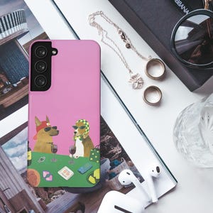 May include: A pink phone case with a cartoon illustration of two dogs wearing sunglasses and drinking wine at a poker table. The image also includes sunglasses, jewellery, rings, a glass, and wireless earbuds.